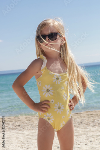Portrait of 4 years old girl in yellow swimsuit and long blonde hair with blue sea and sky on the sandy beach