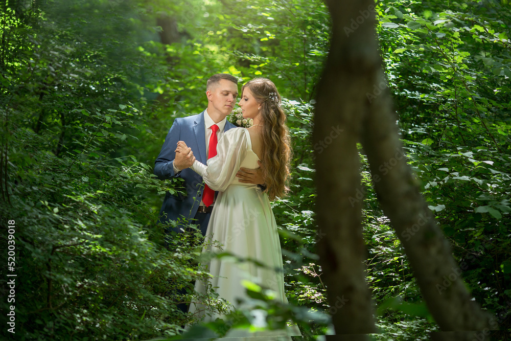 a guy in a suit and a girl in a white dress are standing in the forest and hugging.