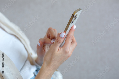 A close-up photo of a beautiful woman s hand with a white manicure is using a gold-colored phone  typing a message on a smartphone