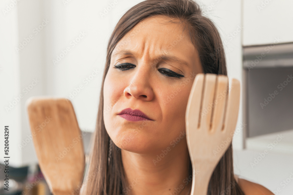 Young brunette woman in her kitchen trying to choose between using a spoon or a wooden fork. Close up image of serious young beautiful woman while holding wooden kitchen utensils with both hands. Young brunette woman in her kitchen trying to choose between using a spoon or a wooden fork. Close up image of serious young beautiful woman while holding wooden kitchen utensils with both hands.