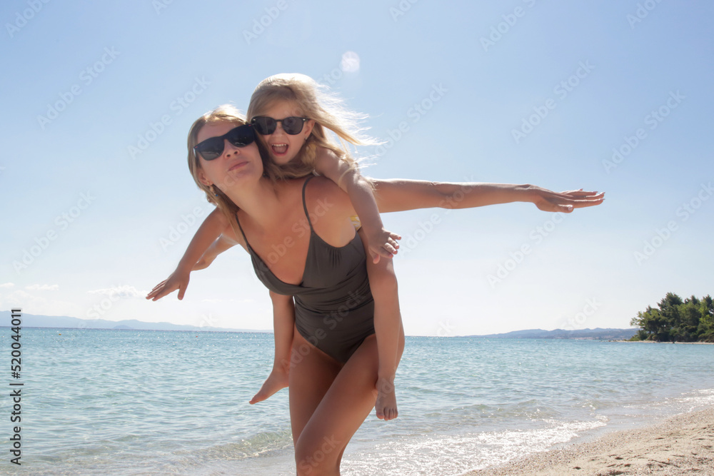 Mother carrying daughter on her back on the beach. Happy idyllic family summer vacation concept. Mother carrying daughter on her back on the beach. Happy idyllic family summer vacation concept.