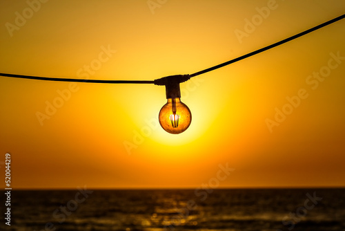 Concept of solar energy. Sun shines trhrough the modern energy saving filament led bulb edison style light lamp against of sky and sea in the sunset. Ecology concept.