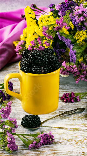 Fresh blackberry berries in a cup and spoon. A yellow cup and dry statice flowers. A bright summer composition.