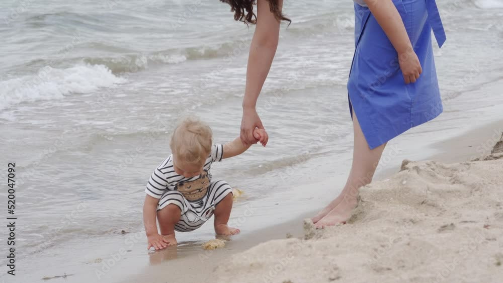 Mother and little cute child are walking on beach, mother and 1.5 year old toddler play near sea waves in summer. The central city beach in the resort town of Evpatoria, Crimea. Mother and little cute child are walking on beach, mother and 1.5 year old toddler play near sea waves in summer. The central city beach in the resort town of Evpatoria, Crimea.