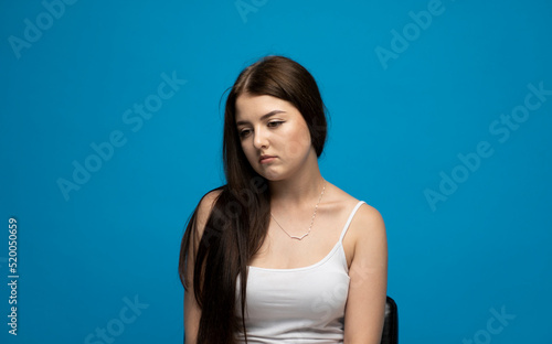 Portrait of upset young beautifil brunette girl with long hair in a white t-shirt standing with puffy cheeks isolated over blue background. Emotion of angry and upset.