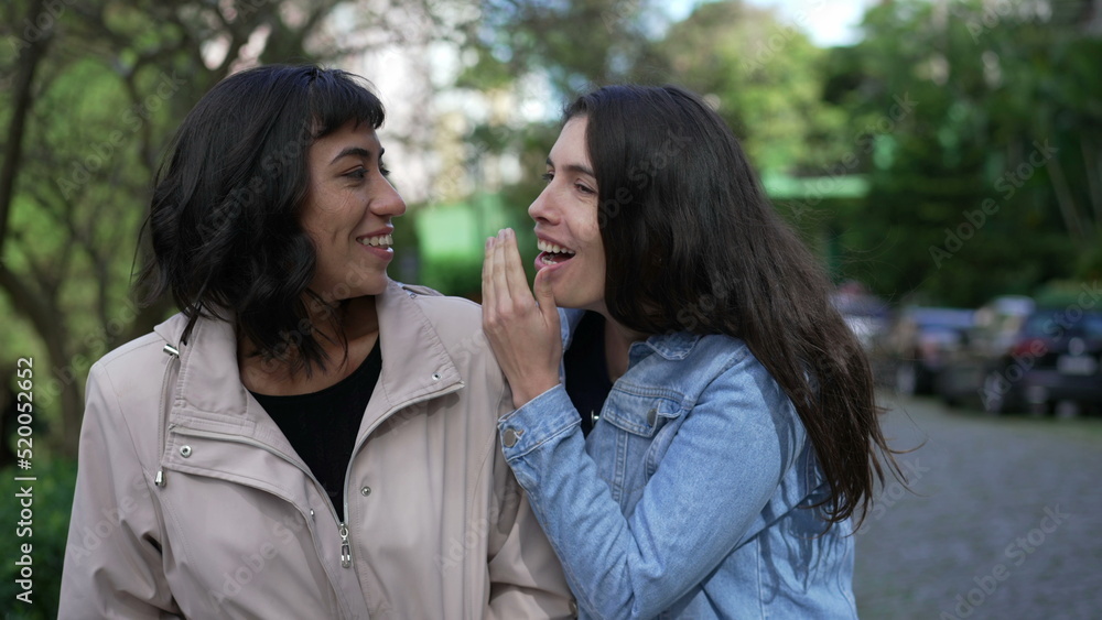 Two female friends gossiping while walking outside in street. Young women conversation on sidewalk Two female friends gossiping while walking outside in street. Young women conversation on sidewalk