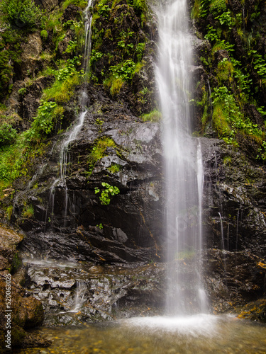 Detalle de una cascada de un rio de montaña que cae entre rocas y vegetación verde.