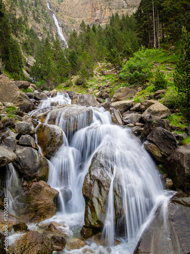 Cascada de alta montaña que recorre el valle entre grandes rocas y rodeado de vegetación y montañas.