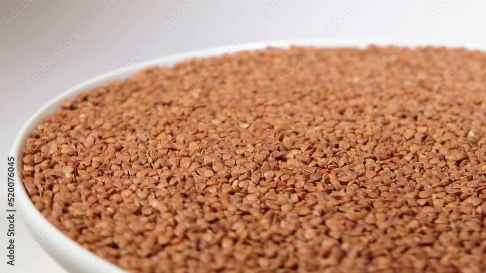 Dry Uncooked Brown Buckwheat Groats on White Plate Rotating against White Background. Raw Large Buckwheat Grains. Russian, Ukrainian and Belarusian Culture