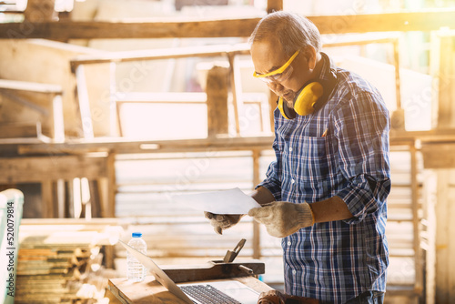 professional senior carpenter using laptop computer for wooden furniture handcraft project working in wood workshop