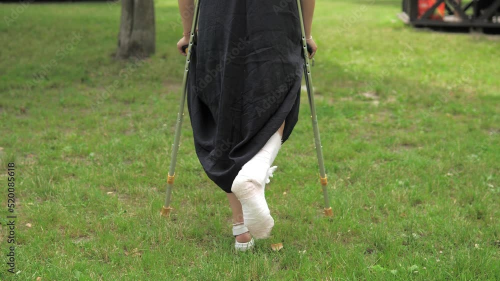 A young girl with a cast walks around the park on crutches on the lawn