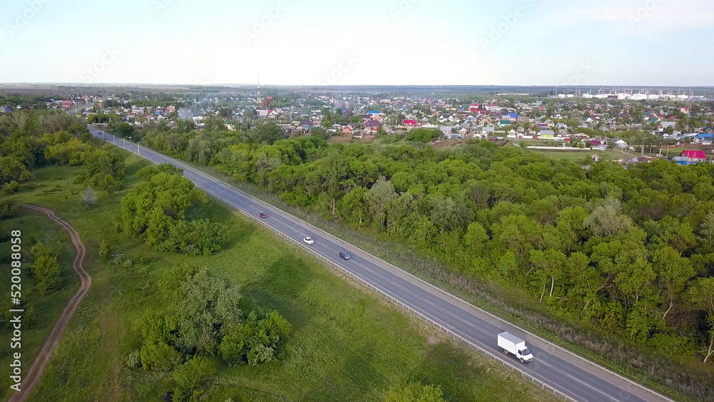 Top view of highway passing through village with forest. Clip. Track with passing cars on background of village with green forest and horizon with sky