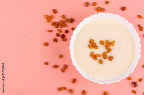 Semolina porridge with raisins in a beautiful white plate (bowl) on a peach-pink background. Place for text, recipe. Contains gluten.