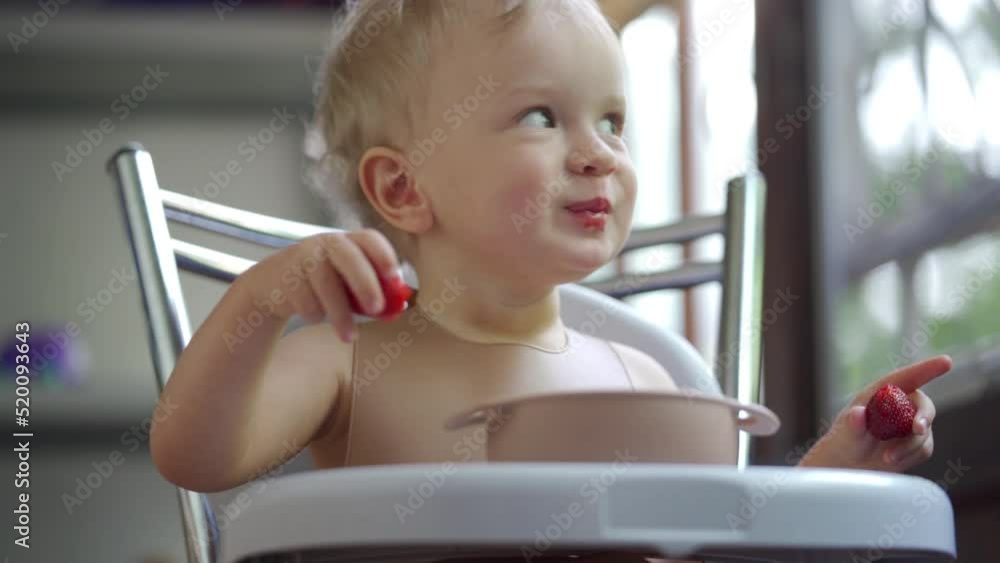 Baby boy eating strawberries for the first time, 1.5 year old toddler child sitting on highchair eats strawberry fruit on the balcony in summer. Baby boy eating strawberries for the first time, 1.5 year old toddler child sitting on highchair eats strawberry fruit on the balcony in summer.
