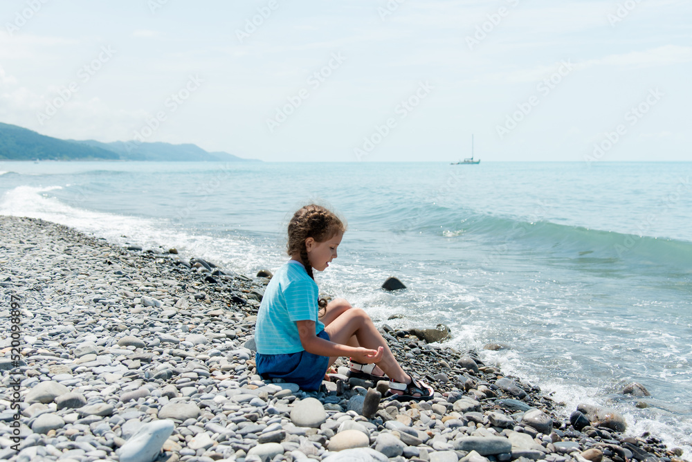 Little cute girl sits on a pebble beach and plays with pebbles