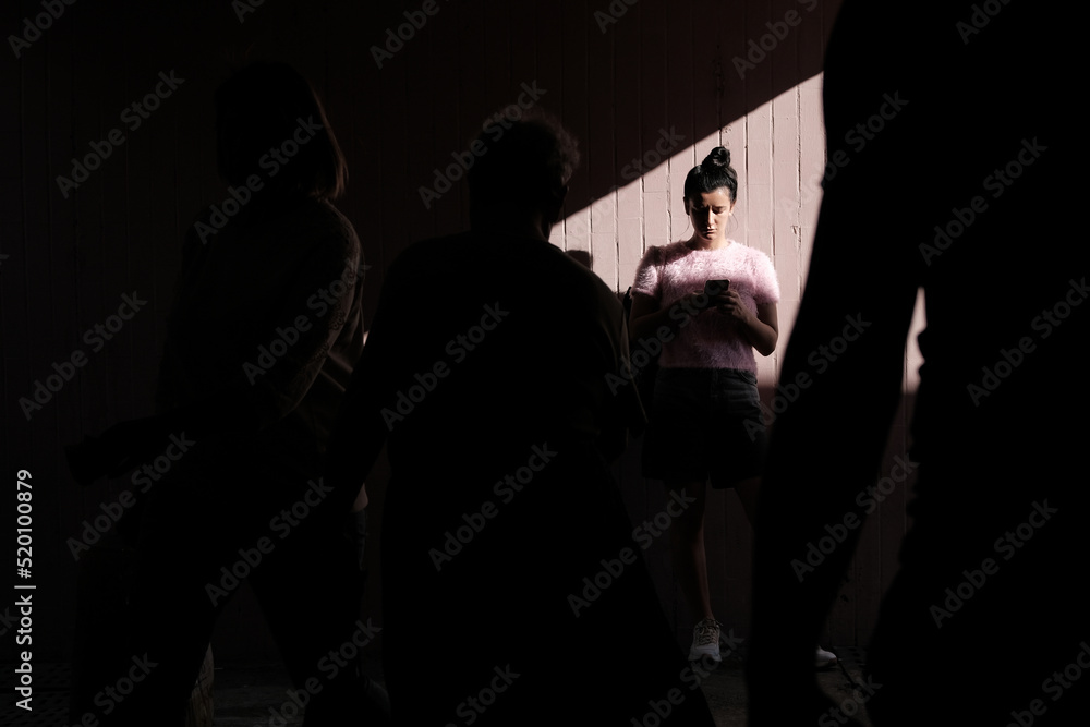 Young woman looking at a phone, standing against wall in underground pedestrian crossing tunnel with contrast shadows and light during sunny day while many people passing by. Street style photography
