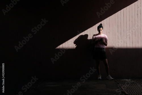 Young woman looking at a phone, standing against wall in underground pedestrian crossing tunnel with contrast shadows and light during sunny summer day. Street style photography