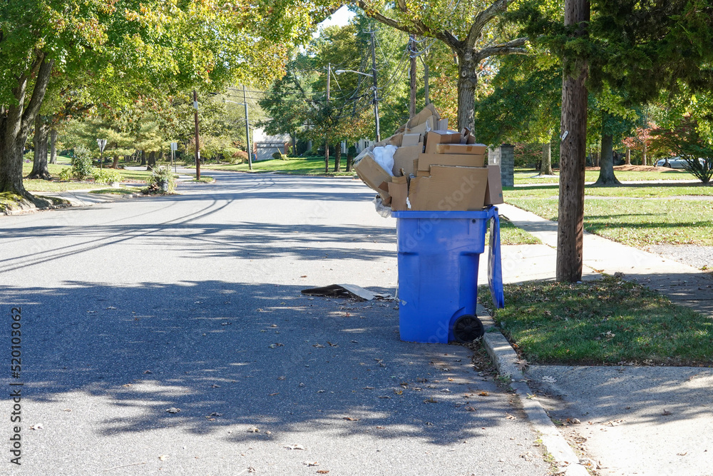 Blue Recycling Bin that is over stuffed with cardboard is seen on a residential street by a curb waiting for pickup Blue Recycling Bin that is over stuffed with cardboard is seen on a residential street by a curb waiting for pickup