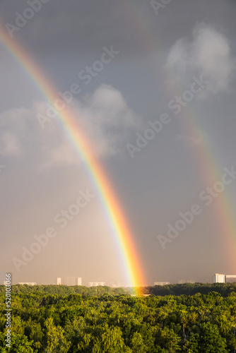 double rainbow in gray blue sky over sunlit forest before thunderstorm on sunny summer day