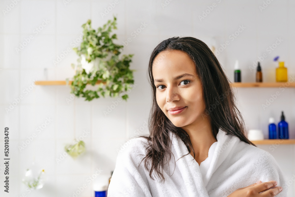 Portrait of young beautiful hispanic woman in white bathrobe with cosmetic bottle on the background. Confident young woman feeling harmonous and calm in her body. Wellness Portrait of young beautiful hispanic woman in white bathrobe with cosmetic bottle on the background. Confident young woman feeling harmonous and calm in her body. Wellness