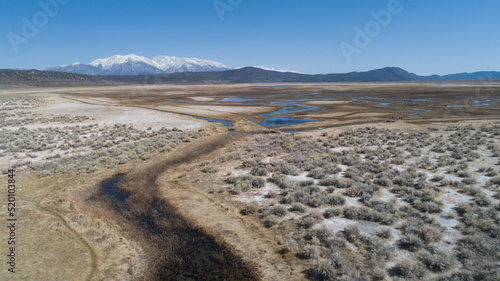 Drone view of the high desert in late spring when the natural springs are flowing making wetlands and salt deposits. Snowcapped mountians in the distance set apart from the stark blue sky