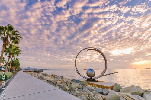 Molos Promenade and skyline of the coast in Limassol city in Cyprus at cloudy sunrise. View of boardwalk pier path landmark with palm trees, sculpture, the Mediterranean sea and people walking.	
