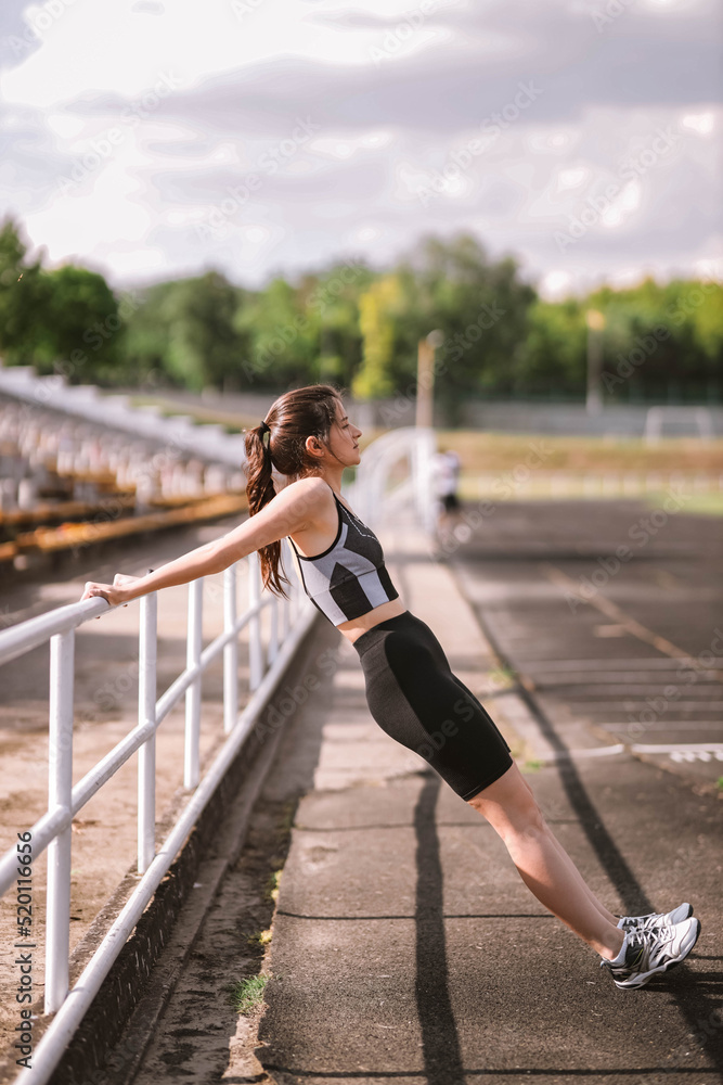 Beautiful Caucasian girl athlete in dressed in sportswear: shorts and top, sneakers with fitness tracker stretching on stadium in summer. Happy female outdoor sport and healthy lifestyle concept