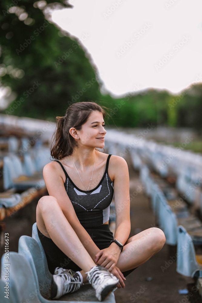 Beautiful Caucasian girl athlete in dressed in sportswear: shorts and top, sneakers with fitness tracker sits in armchair on stadium in summer. Happy female outdoor sport and healthy lifestyle concept