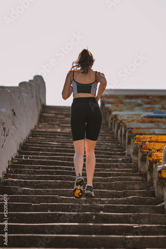 Beautiful Caucasian girl athlete dressed in sportswear: shorts and top, sneakers, fitness tracker runs on stadium stairs in summer. Happy young female outdoor sport, workout, healthy lifestyle concept