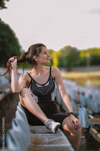 Fototapeta Naklejka Na Ścianę i Meble -  Beautiful Caucasian girl athlete in dressed in sportswear: shorts and top, sneakers with fitness tracker sits in armchair on stadium in summer. Happy female outdoor sport and healthy lifestyle concept