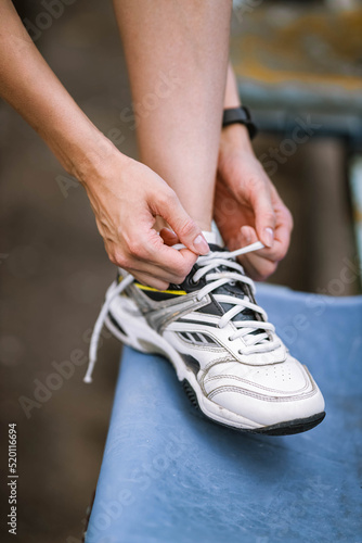 Closeup photo of athlete girl's legs in sneakers, she ties shoes in summer on a stadium chair outside with fitness tracker. Happy young female sport, workout, travelling, healthy lifestyle concept