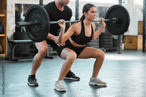 Fototapeta Naklejka Na Ścianę i Meble -  Active woman bodybuilder training and weight lifting with a personal trainer at the gym. Fit and athletic young female athlete lifts a barbell with her coach at a fitness facility