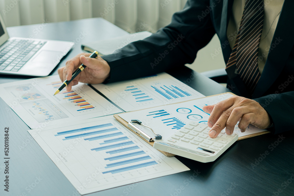 A businessman presses a white calculator to calculate numbers in company financial documents. The finance department prepares accounting documents, analyzing income and expenses in the business. A businessman presses a white calculator to calculate numbers in company financial documents. The finance department prepares accounting documents, analyzing income and expenses in the business.