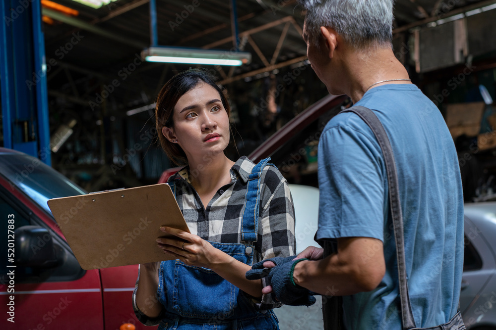 Senior Asian mechanic teaches a female trainee holding a clipboard to write down the list of engines that need to be repaired in the garage. small business and car repair service concept