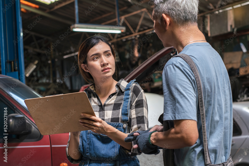 Senior Asian mechanic teaches a female trainee holding a clipboard to write down the list of engines that need to be repaired in the garage. small business and car repair service concept Senior Asian mechanic teaches a female trainee holding a clipboard to write down the list of engines that need to be repaired in the garage. small business and car repair service concept