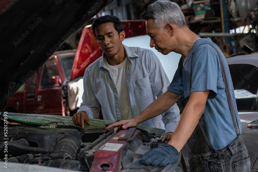 Elderly Asian mechanic standing and talking to a male customer brought a pickup truck to fix it. Talk to inform the information about the broken engine for rent to be repaired in the garage.