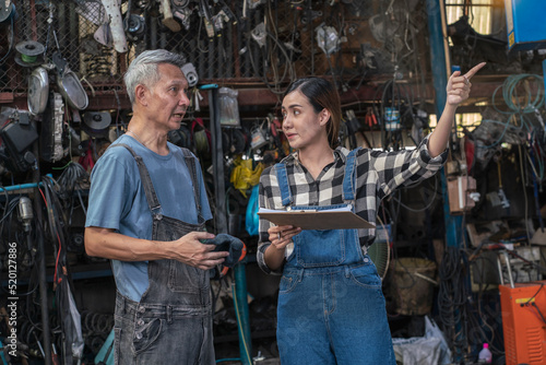 Senior Asian mechanic teaches a female trainee holding a clipboard to write down the list of engines that need to be repaired in the garage. small business and car repair service concept