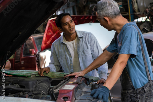 Asian man stands and talks to a senior Asian mechanic overseeing pickup truck engines and looking at repair parts in the garage. Sevices Business concept.