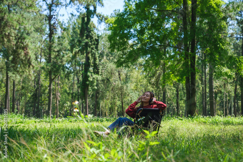 Portrait of a beautiful young asian woman enjoy listening to music with headphone while sitting on a camping chair in the park