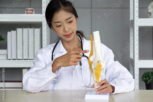 Young asian doctor in white gown and stethoscope pointing at anterior cruciate ligament on deluxe functional knee joint model. photo