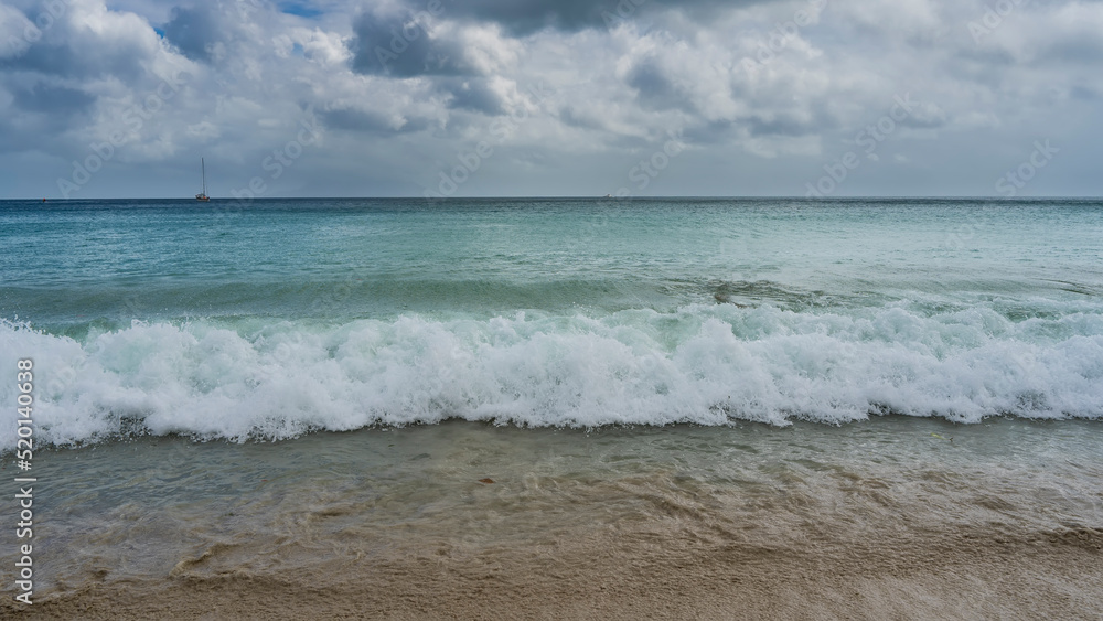 A wave of surf rolls onto the beach. Foam on the sand. In the distance, a tiny ship is visible in the turquoise ocean. Picturesque clouds in the sky. Seychelles. Mahe Island. Beau Vallon Beach