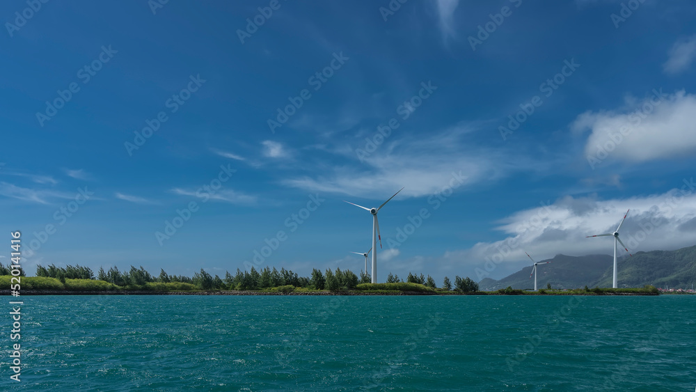 Wind turbines are installed on the island. There is a turquoise ocean all around. A mountain against a blue sky. Seychelles