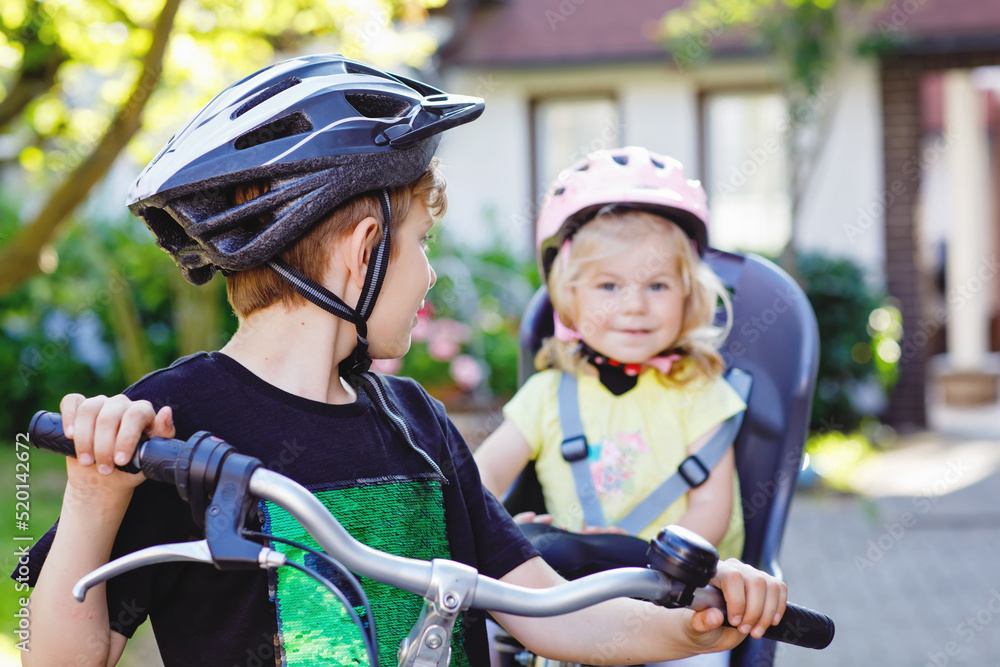 Obraz premium Portrait of little toddler girl with security helmet sitting in bike seat of brother. Kid boy on bicycle with lovely sister. Safe and child protection concept. Family weekend activity trip.