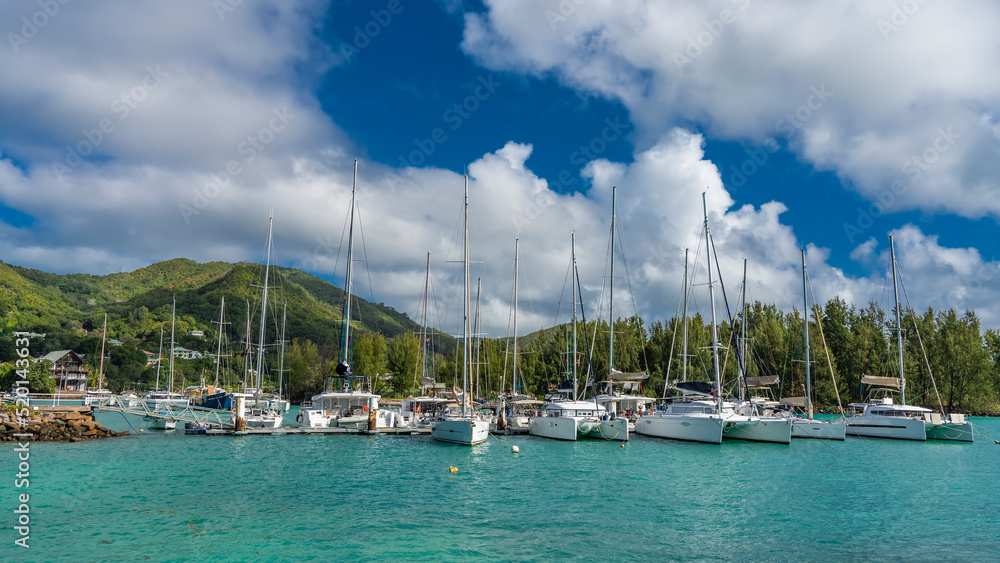 A row of white yachts with masts stand along the pier in the aquamarine ocean. Green mountains against a blue sky with clouds. Seychelles