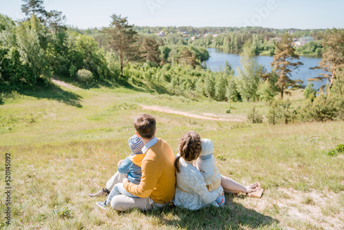 Young family with two children sits on a green hill and looks at the panoramic view of the countryside, the concept of a dream of living in a big country house