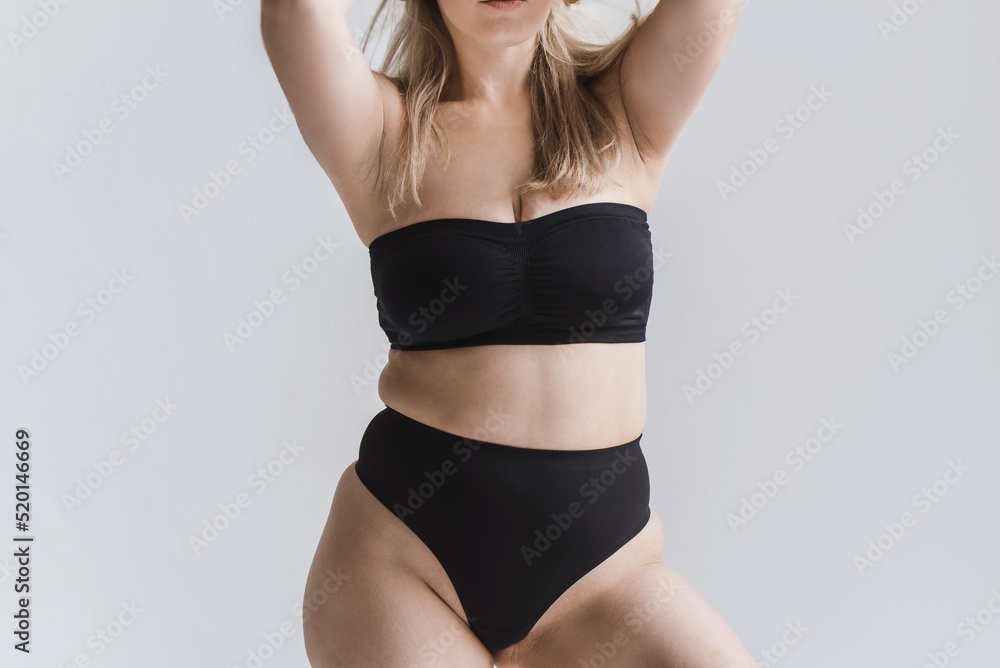 Naklejka premium young overweight woman in sports minimalist black lingerie poses near large window. Plus size model show body. Folds at the waist. Unrecognizable plus size faceless model