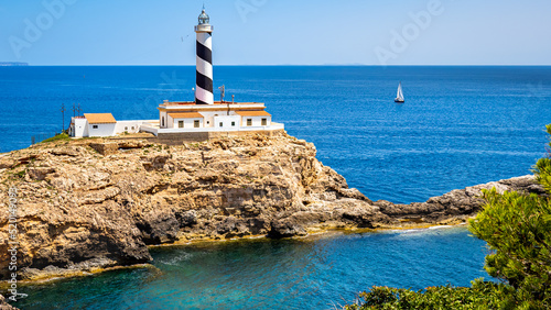 Mallorca bay Es Mular with lighthouse Faro de Cala Figuera on a peninsula in front of a sailboat on the mediterranean sea with cape Cap Blanc and islands of Cabrera national park on the horizon.