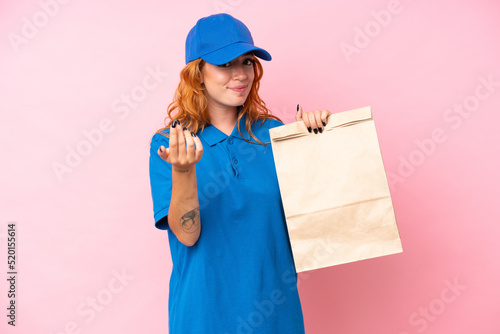 Young caucasian woman taking a bag of takeaway food isolated on pink background inviting to come with hand. Happy that you came