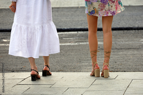 Slim legs of two girls in summer dresses standing on a street. Female fashion in summer city