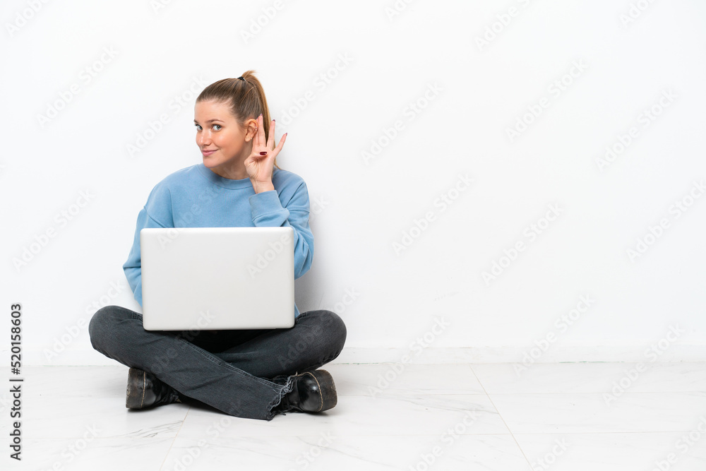 Young woman with a laptop sitting on the floor listening to something by putting hand on the ear Young woman with a laptop sitting on the floor listening to something by putting hand on the ear